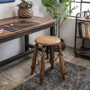 Op de afbeelding: Een houten verstelbare kruk met een ronde zitting en drie schuine poten. De poten zijn verbonden door metalen steunen. De kruk staat op een grijs tapijt, in de buurt van een houten bureau en een metalen plank.