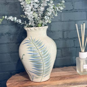 Op de afbeelding: Een crèmekleurige keramische vaas met een gerimpelde rand en een geschilderd blauw en groen bladerontwerp. De vaas bevat kunstmatige eucalyptus takken. De vaas staat op een houten tafel naast een glazen diffuserfles.