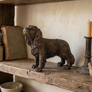 May include: A brown dog figurine with a detailed fur texture stands on a rustic wooden shelf. The dog is looking upwards. A candle and a small bird figurine are also on the shelf.