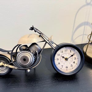 Könnte beinhalten: Eine dekorative Motorradfigur aus Metall mit einem Zifferblatt als Vorderrad. Die Uhr hat ein cremefarbenes Zifferblatt mit schwarzen arabischen Ziffern und schwarzen Zeigern. Der Motorradkörper ist in einem dunklen Metallton gehalten.