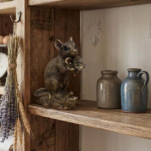 Peut inclure: Une figurine de souris couleur bronze debout sur une base texturée, tenant une fleur dans ses pattes avant. La souris a des yeux sombres et une texture de fourrure détaillée. À côté de la figurine se trouvent deux petits pots en céramique.