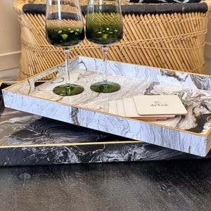 May include: Two rectangular serving trays with marble patterns, one black and gold, the other white and grey with gold trim. Two green-stemmed wine glasses and coasters with the word "drink" are on the top tray. A wicker basket is in the background.