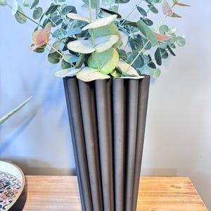 Puede incluir: Un jarrón acanalado gris oscuro contiene ramas de eucalipto con hojas verdes y rosas. El jarrón tiene forma cilíndrica con estrías verticales. El arreglo está sobre una superficie de madera.