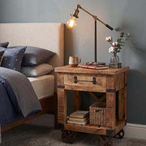 Könnte beinhalten: Ein Nachttisch aus Holz mit einer Schublade, einer unteren Ablage und vier Metallrädern. Der Tisch hat dunkle Metalleckbeschläge und einen Griff. Darauf stehen eine Schreibtischlampe aus Messing, eine Keramiktasse, ein Notizbuch und eine Glasvase.