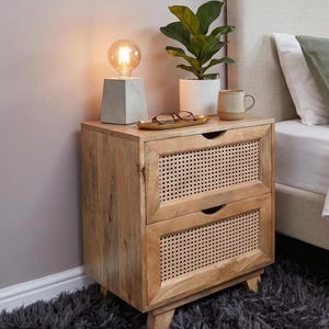 Puede incluir: Una mesita de noche de madera clara con dos cajones que presentan paneles de rejilla tejida y tiradores integrados. La superficie superior sostiene una lámpara con base de hormigón, una planta verde, una taza beige y unas gafas.
