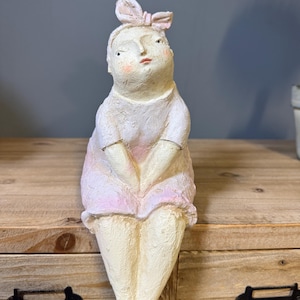 May include: A whimsical, seated figurine with a light pink dress and matching bow. The sculpture has a textured, off-white finish with subtle pink accents on the cheeks and dress. The figurine is posed with hands clasped and legs crossed.
