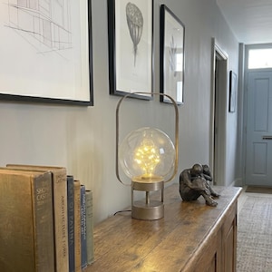 Könnte beinhalten: Eine dekorative Lampe mit einer durchsichtigen Glaskugel, die mit warmweißen Lichtern gefüllt ist, steht auf einem Holzschrank. Die Lampe hat einen Metallsockel und -rahmen. Gerahmte botanische Drucke und Bücher befinden sich ebenfalls auf dem Schrank.