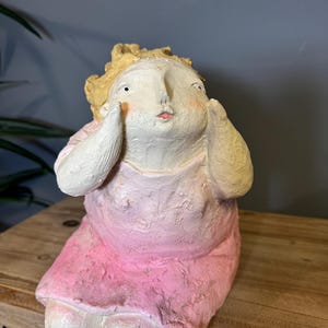 Pode incluir: Uma figura caprichosa pintada à mão de uma pessoa sentada com as mãos perto do rosto. A figura tem uma tez clara, um vestido rosa e cabelo loiro. A escultura está sobre uma superfície de madeira.
