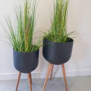 Può includere: Due vasi neri con erba alta e verde e gambe in legno. I vasi hanno una forma arrotondata e sono sollevati da tre gambe in legno marrone chiaro angolate. L'erba ha accenti gialli e marroni.