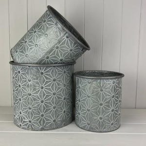 May include: Three galvanized metal plant pots in different sizes. The pots are a muted grey colour and feature a repeating geometric starburst pattern. The largest pot is approximately 25 cm in diameter and 20 cm tall. The smallest pot is about 15 cm in diameter and 15 cm tall.