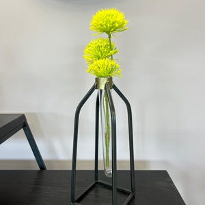 May include: A black metal vase with a clear glass tube in the center. The vase has a square base and a rectangular frame. The vase is filled with a single stem of yellow flowers.