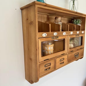 Peut inclure: Organisateur mural en bois avec plusieurs compartiments et tiroirs. Comprend des sections vitrées et des étiquettes numérotées. La finition en bois naturel et la quincaillerie de style antique lui confèrent un charme rustique. Contient de petits objets décoratifs.