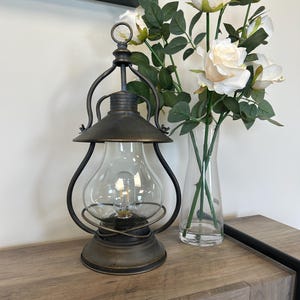 Op de afbeelding: Een antieke lantaarn met een heldere glazen lamp en een donker metalen frame. De lantaarn staat op een houten oppervlak naast een vaas met witte rozen. De lantaarn heeft een decoratieve bovenring.