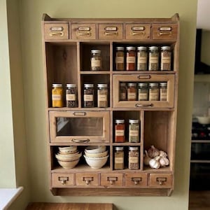 Può includere: Una spezieria da parete in legno con cinque cassetti superiori con maniglie in ottone, diversi ripiani aperti, due armadietti con frontale in vetro e cinque cassetti inferiori. Contiene vari barattoli di spezie, ciotole in ceramica e aglio.
