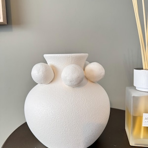 May include: A white ceramic vase with a textured surface and three spherical accents. The vase has a rounded body and a flared opening. The vase is displayed on a dark brown surface.