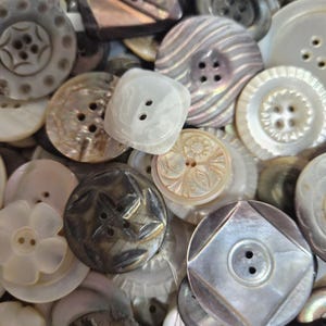May include: A collection of various vintage buttons in shades of white, gray, and brown. The buttons are made of different materials, including mother-of-pearl, and feature a variety of shapes, sizes, and designs, such as floral and geometric patterns.