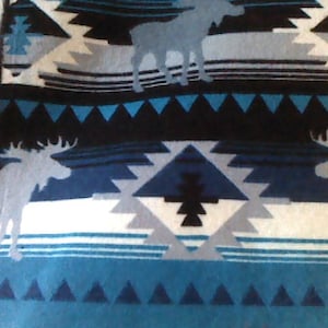 Peut inclure: Une couverture à motifs bleus, noirs et blancs avec un motif géométrique et des silhouettes d'orignaux. La couverture a une bordure à franges et est faite d'un matériau doux. Le motif comprend des triangles et des rayures horizontales.