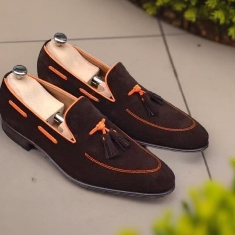 May include: A pair of brown suede loafers with orange stitching and tassels. The loafers have a pointed toe and a leather sole.