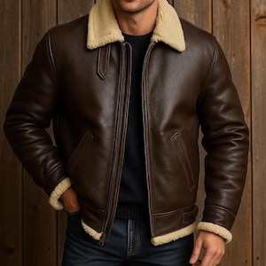 May include: A brown leather jacket with a cream shearling collar and lining. The jacket has a zip-up front, two front pockets, and shearling trim on the cuffs and hem. The jacket is worn over a dark sweater and blue jeans.