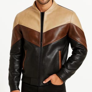 May include: A multi-tone leather jacket featuring a color block design. The jacket has a tan upper section, a brown chevron, and a black lower section. It has a full-length zipper, a stand-up collar, and two side pockets. The jacket is a bomber style.