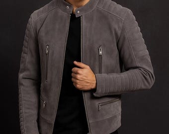 Può includere: Una giacca in camoscio grigio con colletto alla coreana e chiusura a bottone automatico, zip frontale integrale e quattro tasche con cerniera. Le spalle e le maniche presentano un'imbottitura orizzontale trapuntata sopra una maglia nera.