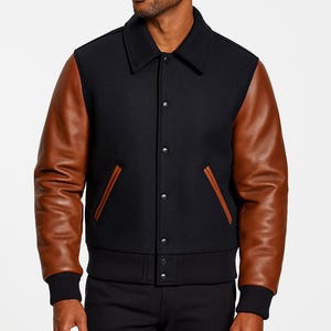 Könnte beinhalten: Schwarze Collegejacke mit braunen Lederärmeln. Die Jacke hat eine Knopfleiste, einen gerippten Saum und Bündchen sowie zwei Vordertaschen mit brauner Einfassung. Der Kragen ist klassisch.