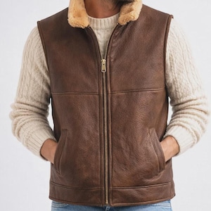 Può includere: Un gilet in pelle marrone con colletto in montone beige e cerniera a tutta lunghezza. Il gilet ha due tasche anteriori e un aspetto strutturato. Il modello indossa un maglione color crema e jeans blu.