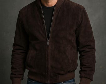 May include: A dark brown suede bomber jacket with a zip-up front, ribbed cuffs, and a ribbed hem. The jacket has two front pockets and a collarless design. The model is wearing a black shirt underneath.