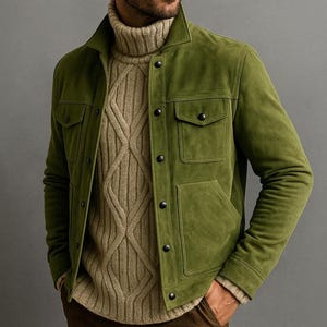May include: A green suede jacket with four front pockets and black buttons. The jacket is worn over a tan cable-knit turtleneck sweater. The outfit is completed with brown trousers. The background is a solid grey.