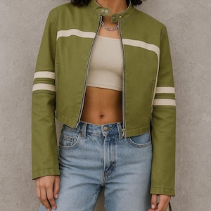 May include: Olive green cropped jacket with a cream-colored stripe across the chest and on the sleeves. The jacket has a stand-up collar, a silver zipper, and two snap closures at the neck. The model is wearing light blue jeans.