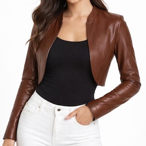 May include: A brown leather bolero jacket with long sleeves. The jacket has a collarless design and a cropped length, revealing a black top and white trousers. The leather appears smooth and has a slight sheen.