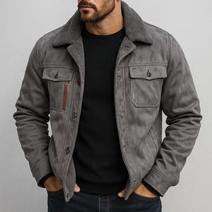 May include: A gray suede jacket with a shearling collar and two chest pockets. The jacket has a button-up front and a brown zipper detail on the left chest pocket. The model is wearing a black sweater underneath.
