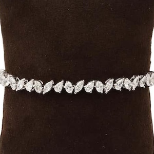 May include: A silver bracelet with a series of pear-shaped, clear gemstones. The bracelet is displayed on a dark brown velvet pillow, creating a contrast that highlights the jewelry's sparkle and design.