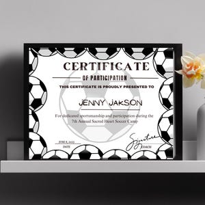 Puede incluir: Certificado de participación enmarcado en negro para Jenny Jakson del 7º Campamento Anual de Fútbol Sacred Heart. El certificado tiene un diseño de balón de fútbol y la fecha 8 de junio de 2023.