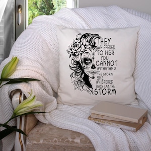 May include: A white throw pillow with a black and white illustration of a woman with a sugar skull face and flowers. The text on the pillow reads "They whispered to her you cannot withstand the storm she whispered back I am the storm."