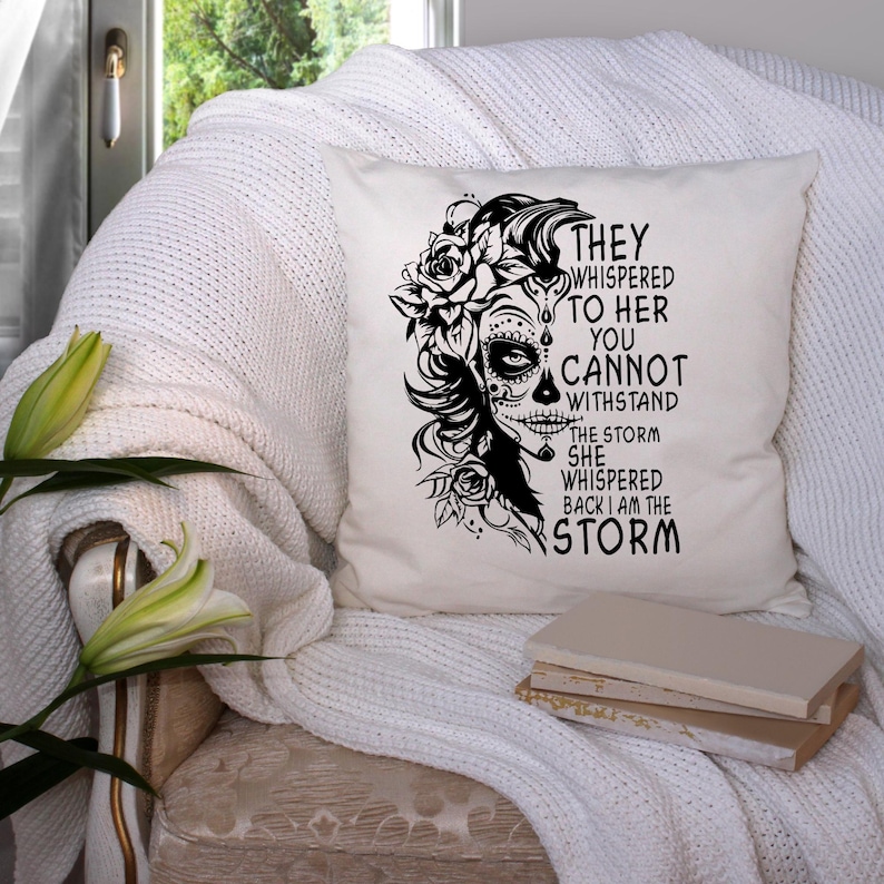 May include: A white throw pillow with a black and white illustration of a woman with a sugar skull face and flowers. The text on the pillow reads "They whispered to her you cannot withstand the storm she whispered back I am the storm."