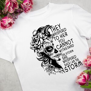 May include: Black and white graphic design featuring a woman with a sugar skull face and flowers. The text reads "They whispered to her you cannot withstand the storm she whispered back I am the storm."
