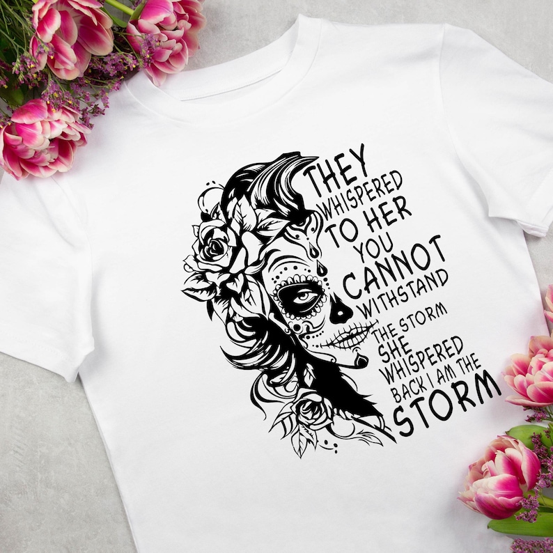 May include: Black and white graphic design featuring a woman with a sugar skull face and flowers. The text reads "They whispered to her you cannot withstand the storm she whispered back I am the storm."