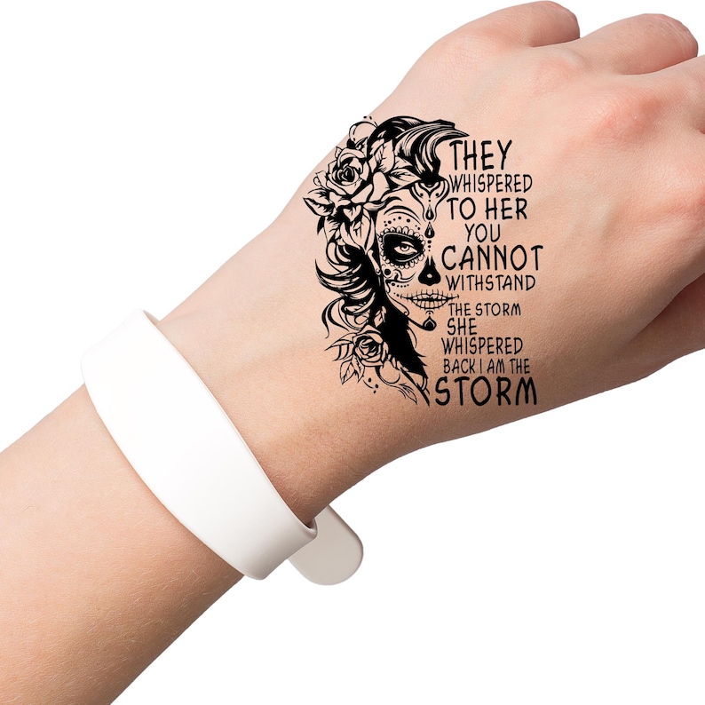 May include: Black and white temporary tattoo with a sugar skull design and the text "They whispered to her you cannot withstand the storm she whispered back I am the storm".
