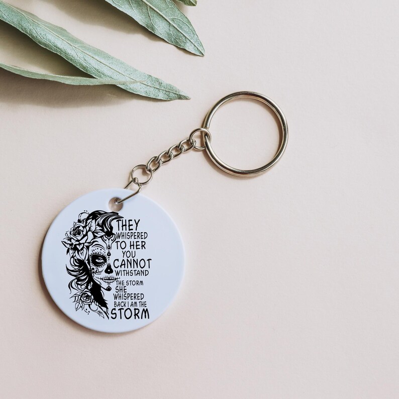 May include: White round keychain with a black and white image of a woman with flowers in her hair and the text "They whispered to her you cannot withstand the storm she whispered back I am the storm".