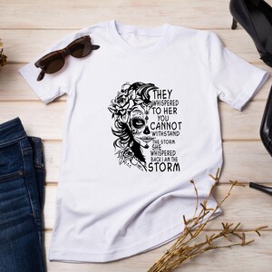 May include: White t-shirt with a black and white graphic of a woman with flowers in her hair and the text "They whispered to her you cannot withstand the storm she whispered back I am the storm."