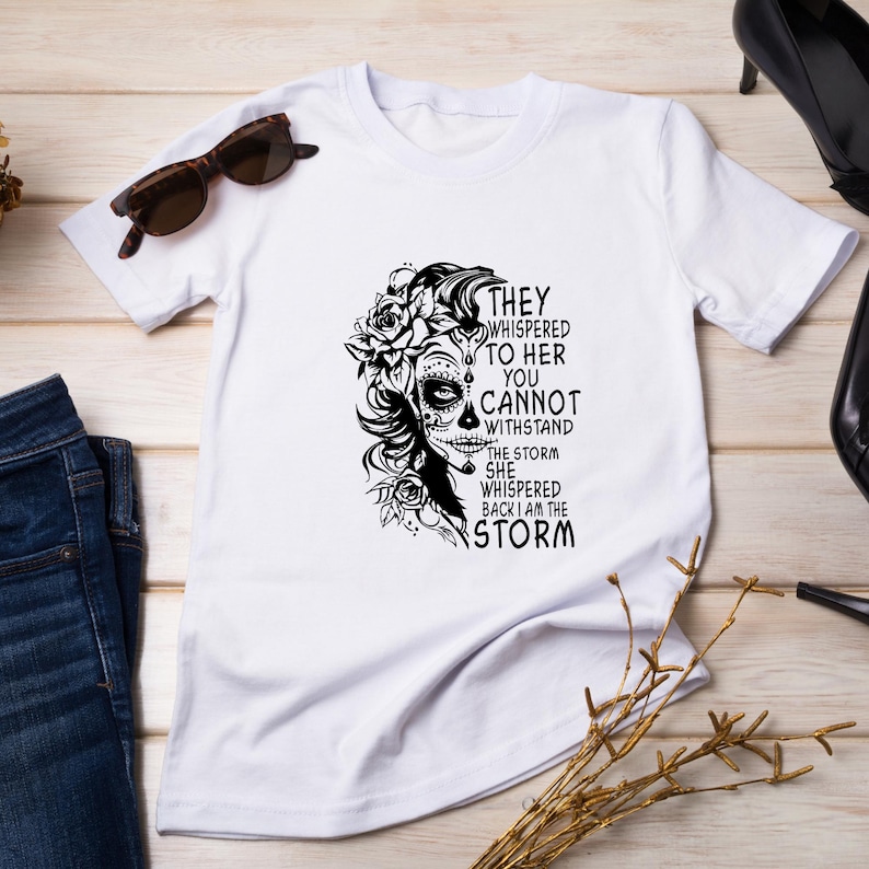 May include: White t-shirt with a black and white graphic of a woman with flowers in her hair and the text "They whispered to her you cannot withstand the storm she whispered back I am the storm."