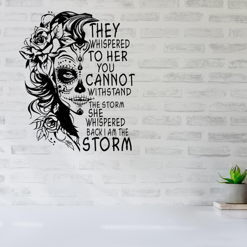May include: Black and white graphic design featuring a woman with a sugar skull face and floral accents. The text reads "They whispered to her you cannot withstand the storm she whispered back I am the storm."