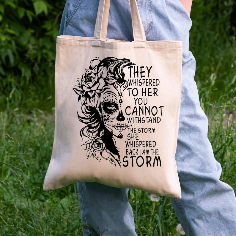 May include: A beige tote bag with a black design of a woman with a sugar skull face and flowers. The text on the bag reads "They whispered to her you cannot withstand the storm she whispered back I am the storm."