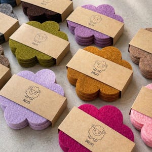 Könnte beinhalten: Eine Sammlung von Filzuntersetzern in verschiedenen Farben, darunter Grün, Lila und Orange. Jeder Untersetzer ist blütenförmig und in einer kleinen Pappschachtel verpackt. Die Schachteln sind mit dem Wort "DOLLYS" und einer Schafillustration versehen.