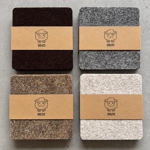 May include: Four square felt coasters in dark brown, grey, light brown, and off-white. Each coaster is wrapped in a tan paper band featuring a black line drawing of a sheep and the text DOLLYS printed in a simple sans-serif font.
