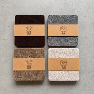 May include: Four square felt coasters in dark brown, grey, tan, and off-white. Each coaster is wrapped in a tan paper band featuring a black line drawing of a sheep and the text DOLLYS. The coasters have a textured, fibrous surface appearance.