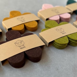Op de afbeelding: Een verzameling gekleurde vilten schoenzolen in verschillende tinten, waaronder bruin, roze, groen en goud. Elke set is verpakt in een kleine kartonnen doos met een schapenlogo en het woord "DOLLYS".