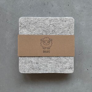 May include: A square light gray felt coaster wrapped in a brown paper band. The band features a black line drawing of a sheep and the text DOLLYS printed in black. The coaster has a textured, fibrous surface and rounded corners.