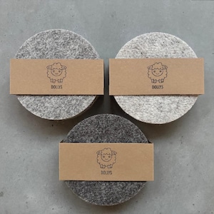 May include: Three circular felt coasters in varying shades of gray, ranging from light to dark. Each coaster is wrapped in a brown paper band featuring a black line drawing of a sheep and the text DOLLYS printed below the illustration.